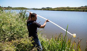 person draws sample water from a sizeable river with a long pole